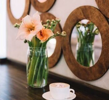 A Flower Vase and a Cup of coffee kept on a table with wooden circular designs on the wall in the background